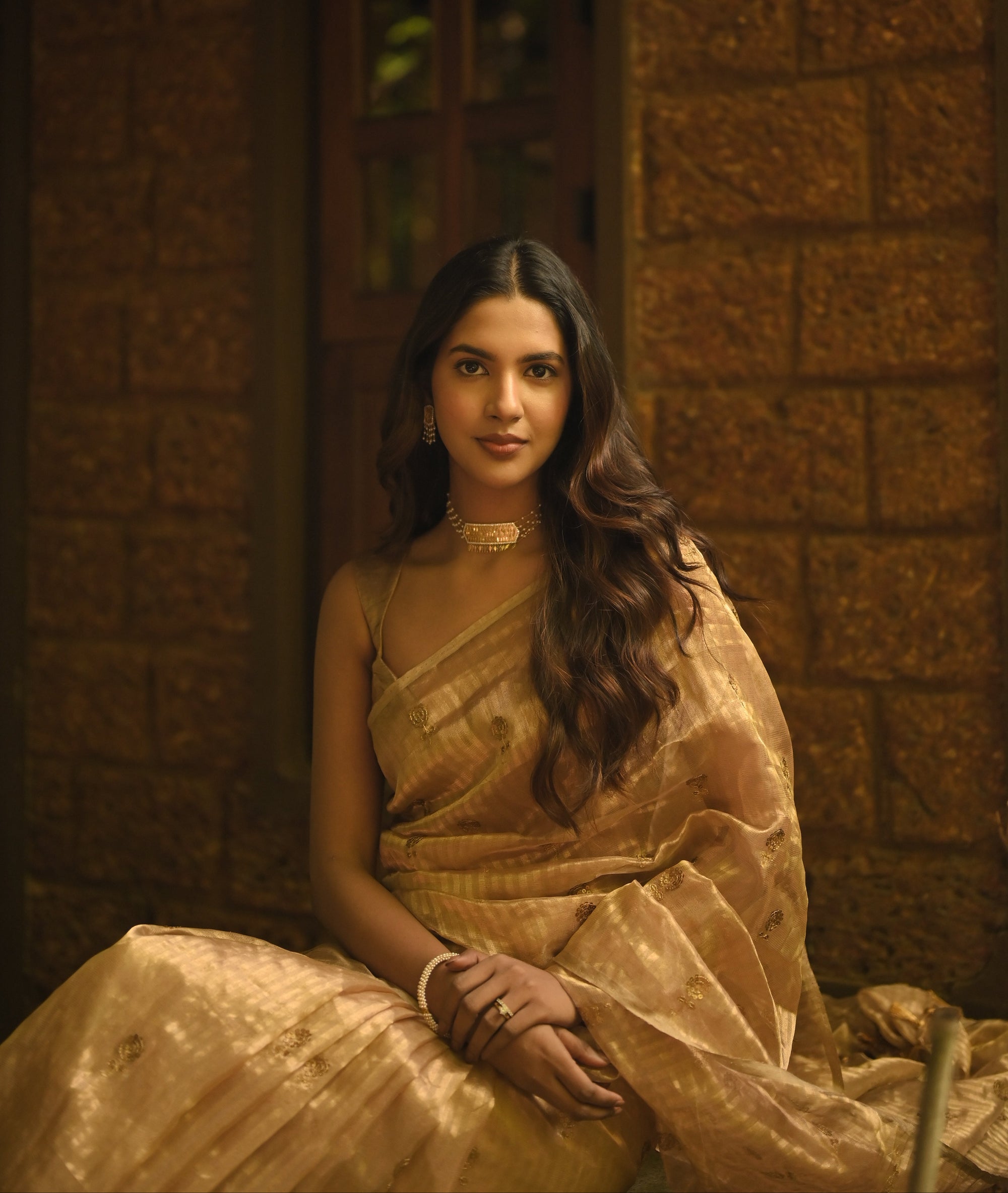 Woman in a gold tissue silk saree sitting against a textured wall.